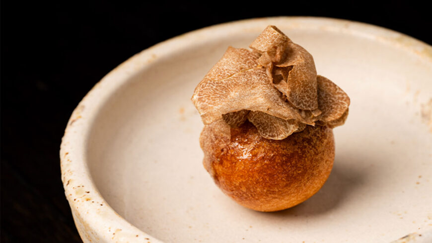 A golden-brown fried potato croquette topped with thinly shaved truffle slices, served on a small off-white ceramic plate against a black background.