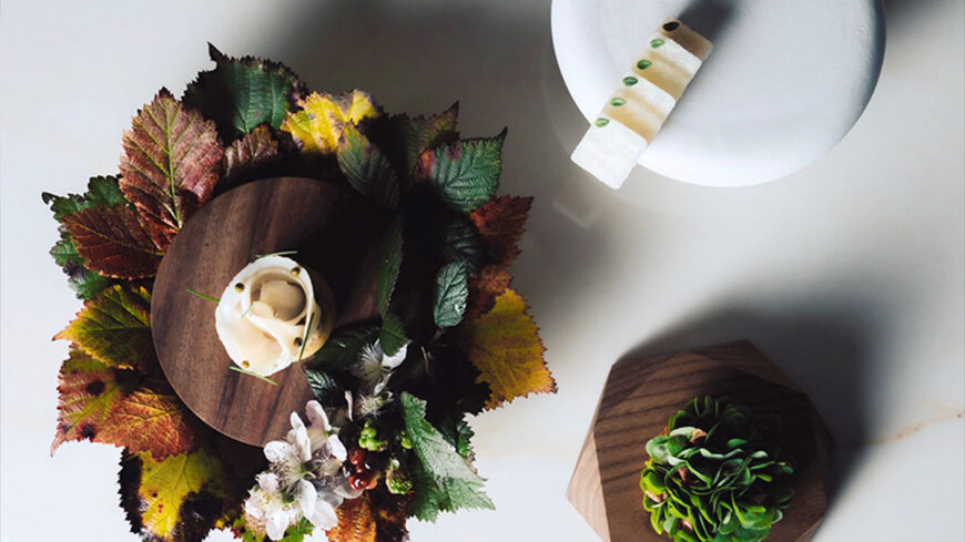 Overhead view of an artistic plated dessert on a wooden serving board surrounded by colorful autumn leaves, with a second minimalist dessert on a white plate nearby.