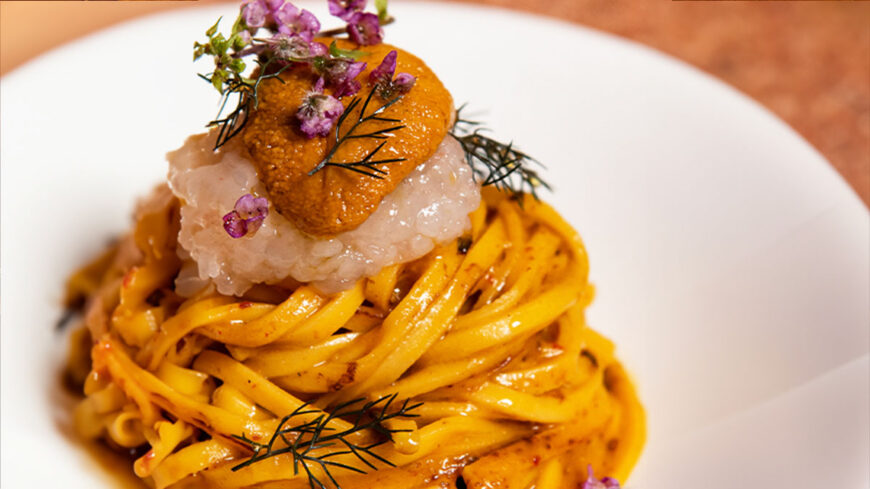Close-up of a nest of fresh pasta topped with creamy sea urchin, translucent seafood, edible flowers, and fresh herbs, served on a white plate.