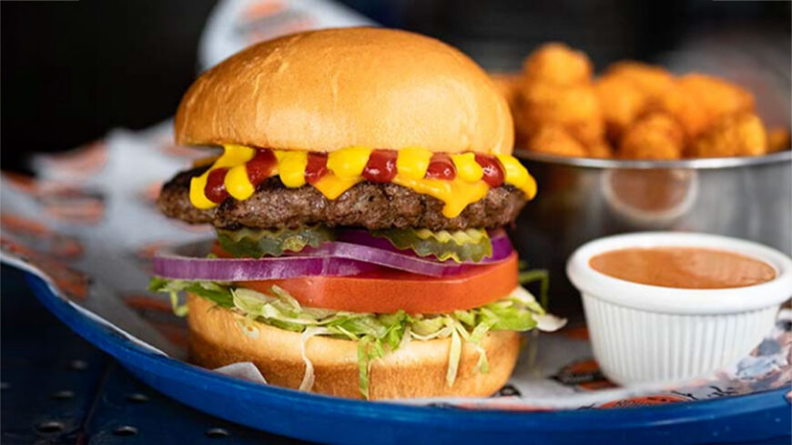 Cheeseburger topped with lettuce, tomato, red onion, pickles, ketchup, and mustard on a brioche bun, served with tater tots and a side of dipping sauce.