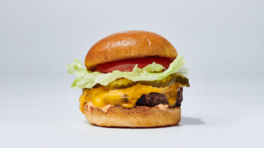 Classic cheeseburger with melted cheddar, lettuce, tomato, pickles, and burger sauce on a toasted bun against a light gray background.