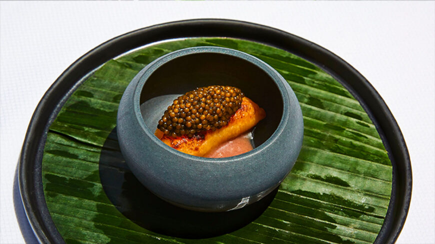 Small black bowl containing a gourmet bite topped with caviar, presented on a large black plate lined with a glossy green leaf.
