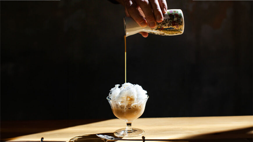 A hand pours a caramel-colored sauce from a decorative ceramic pitcher over a bowl of finely shaved ice in a glass dessert dish, illuminated by warm, dramatic lighting.