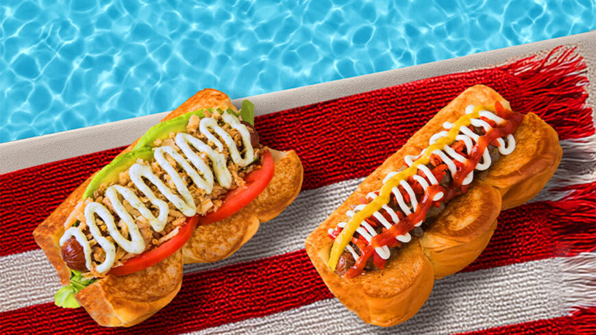 Two specialty hot dogs on buns displayed on a red, white, and blue towel beside a swimming pool. One hot dog is topped with ketchup, mustard, and mayonnaise; the other is topped with lettuce, tomato, crispy onions, and a white sauce.