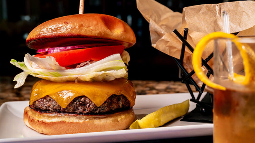 Close-up of a cheeseburger with lettuce, tomato, red onion, and a glossy bun, served with a pickle spear and a drink in the foreground.