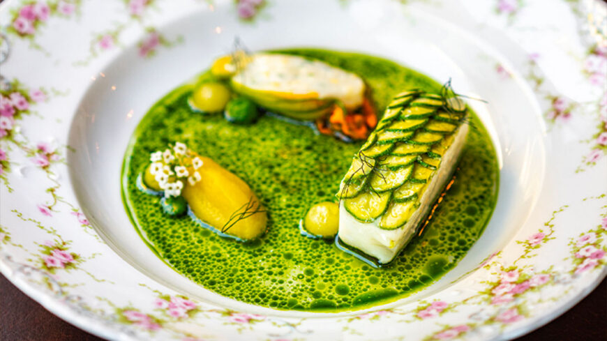 A refined seafood dish featuring a rectangular fillet topped with thin overlapping green vegetable slices, served in a vibrant green herb broth with vegetables and edible flowers on a floral-patterned plate.