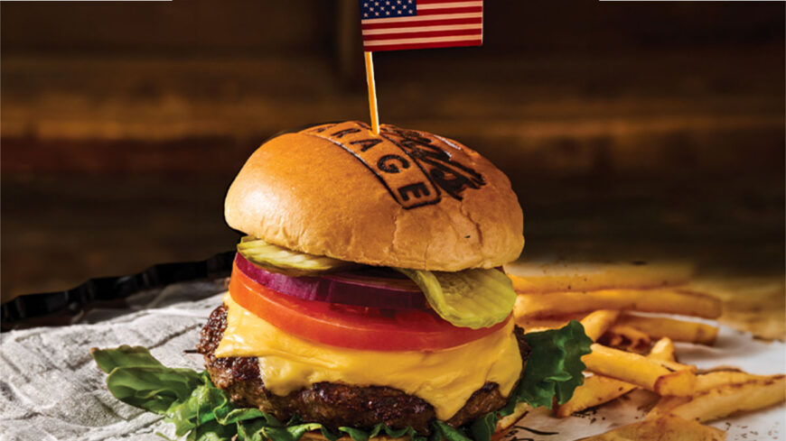Cheeseburger topped with lettuce, tomato, onion, and pickles on a branded bun with a small American flag toothpick, served with French fries.