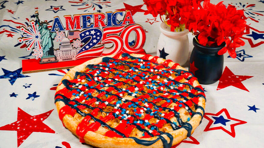 A dessert pizza decorated with red, white, and blue icing and star-shaped sprinkles on a patriotic table setting with flowers and an "America 250" decoration.