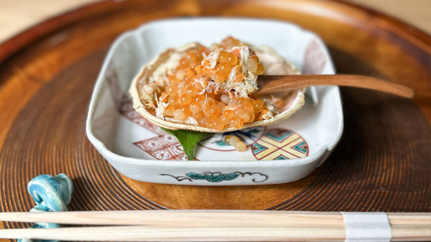 A crab shell filled with crab meat and salmon roe is served on a decorative ceramic plate with a wooden spoon, chopsticks, and a traditional Japanese-style table setting.