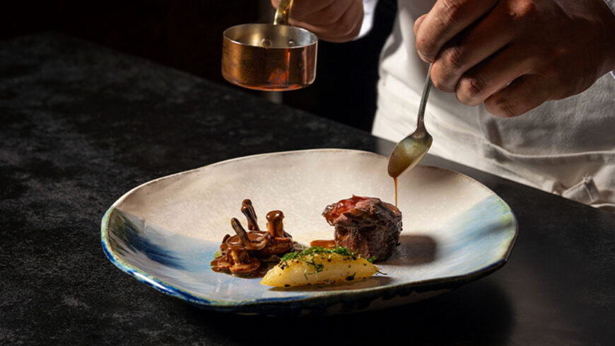 A chef spoons glossy sauce over a seared piece of beef plated with mushrooms and a quenelle of mashed potatoes on a large ceramic dish.