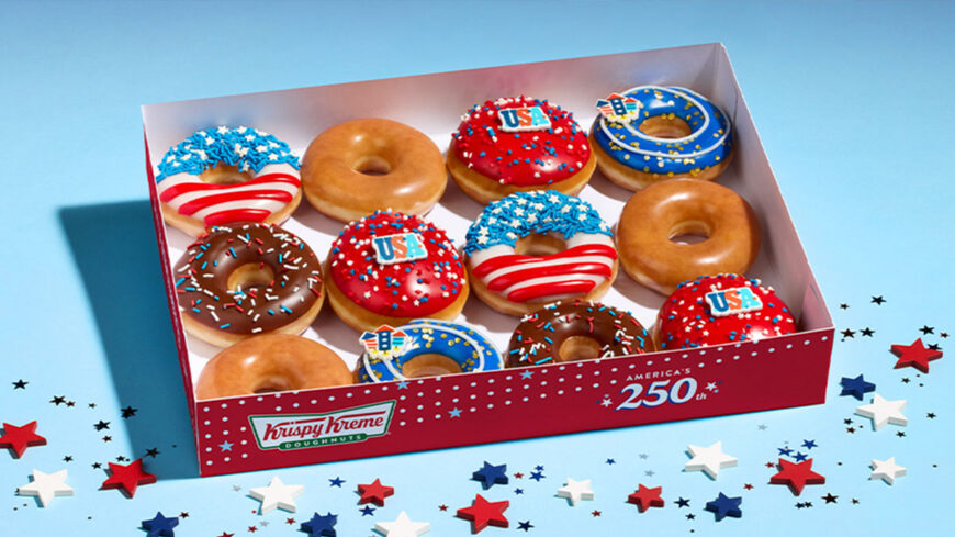 Box of assorted patriotic-themed doughnuts featuring red, white, and blue icing, sprinkles, and USA decorations alongside classic glazed and chocolate sprinkle doughnuts.