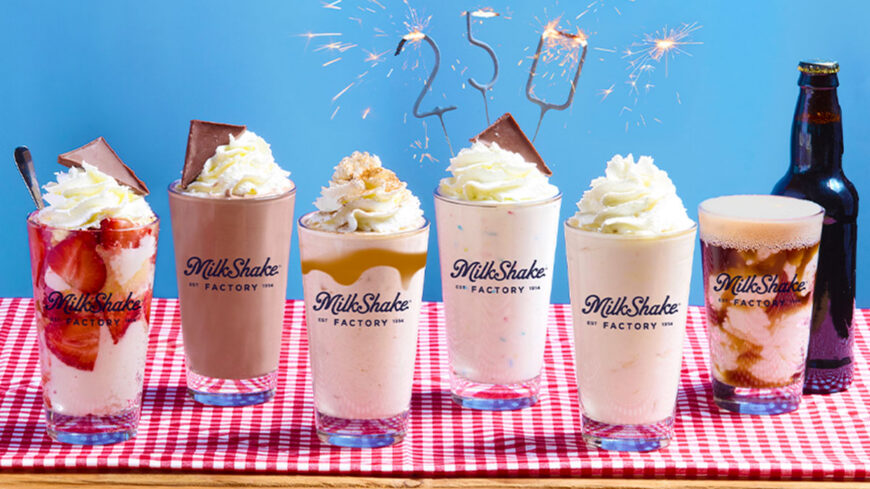 A lineup of six colorful milkshakes topped with whipped cream sits on a red-and-white checkered tablecloth against a blue background. Sparkler candles spelling "250" and a bottle of root beer celebrate a special anniversary.
