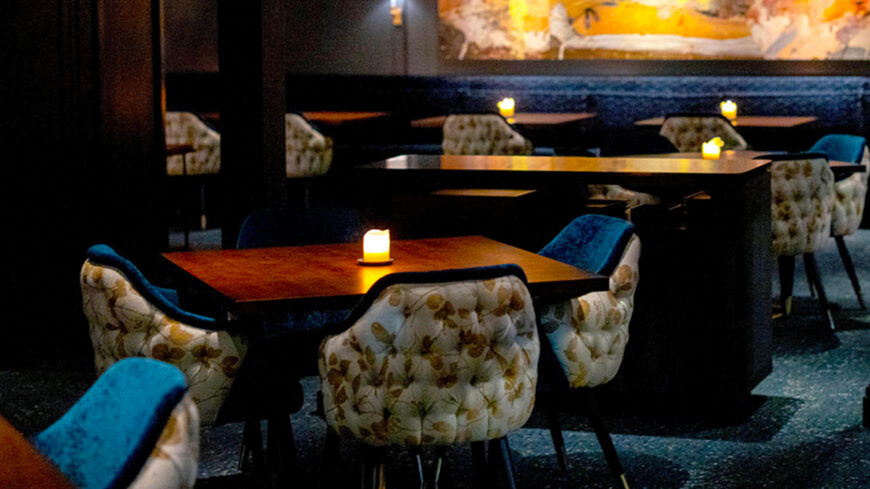 A dimly lit restaurant dining room with candlelit wooden tables and upholstered chairs, creating an intimate, upscale atmosphere.