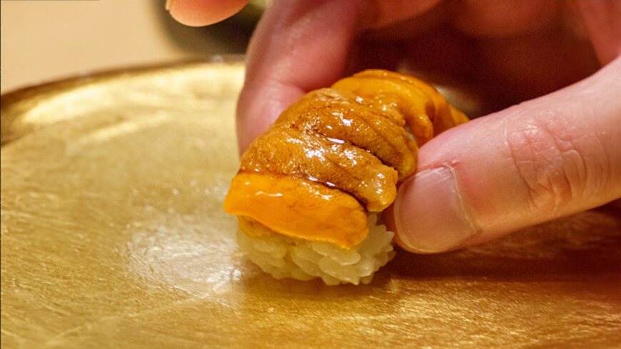 A hand places a piece of sea urchin (uni) nigiri on a gold-colored serving surface, highlighting the sushi's delicate presentation.