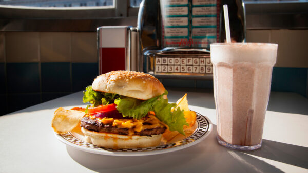 Cheeseburger topped with lettuce, tomato, and onion on a plate with potato chips, served beside a tall milkshake in a retro diner booth.