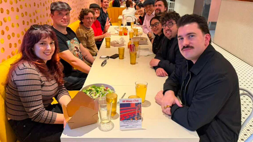 About a dozen people seated around a long table in a brightly colored restaurant, smiling toward the camera during a group dinner. Drinks, plates, and a salad are visible on the table.