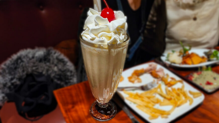 Close-up of a vanilla milkshake topped with whipped cream and a cherry, served in a tall glass on a restaurant table with fries and plates blurred in the background.