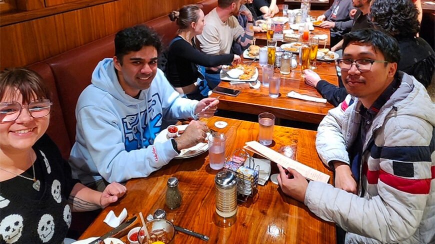 Several diners seated around a wooden table in a restaurant booth, eating and talking during a group meal. Menus, drinks, and plates of food are visible on the table.