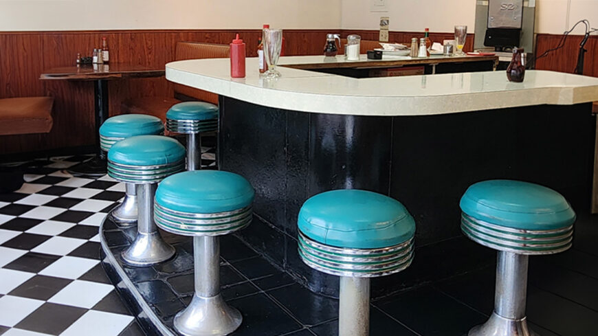 Retro diner counter with turquoise stools, black-and-white checkered floor, and condiment bottles arranged on the countertop.