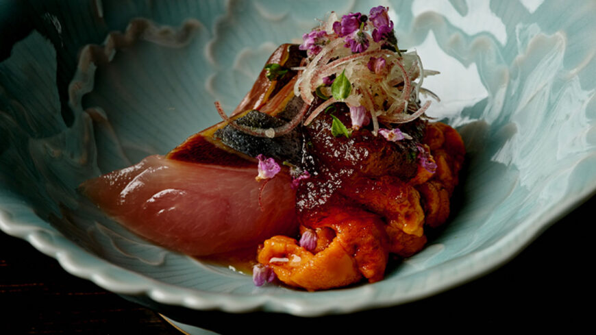 A close-up of an artfully plated seafood dish in a pale blue ceramic bowl, featuring slices of raw fish, sea urchin, glazed shellfish, charred vegetables, and a garnish of thinly sliced onions and small purple edible flowers.