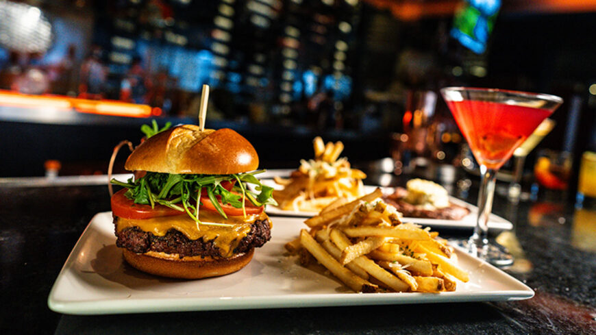 A gourmet cheeseburger topped with lettuce, tomato, and cheese is served with French fries on a white plate. A red cocktail and additional entrées are visible in the softly blurred background of a restaurant.