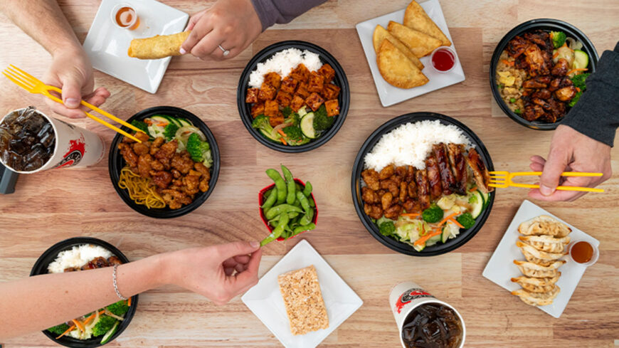Overhead view of a wooden table with several Asian takeout meals, including rice bowls with grilled and fried chicken, noodles, vegetables, dumplings, egg rolls, edamame, crispy treats, and soft drinks. Multiple people are reaching in with chopsticks and hands to share the food.