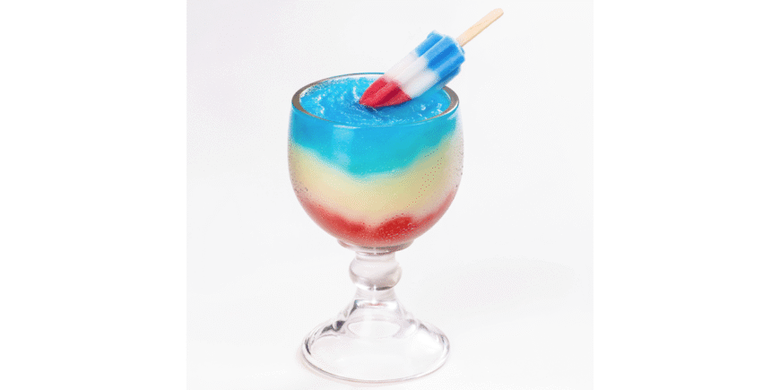 A layered red, white, and blue frozen drink in a stemmed glass, topped with a red, white, and blue popsicle resting on the rim.