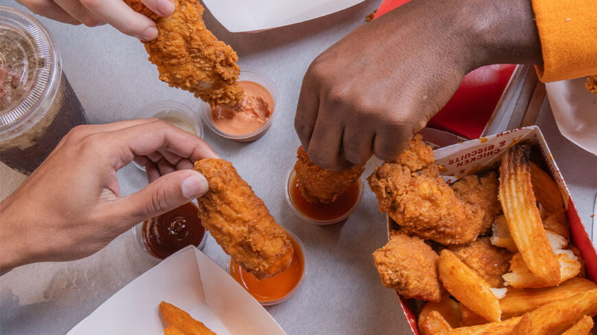 Hands dipping crispy fried chicken wings into various sauces, with seasoned potato wedges and drinks on a shared table.