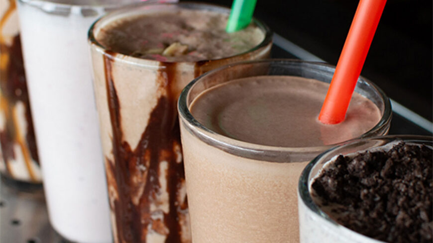 Three milkshakes in glass cups with colorful straws, including chocolate and vanilla flavors, lined up on a counter.