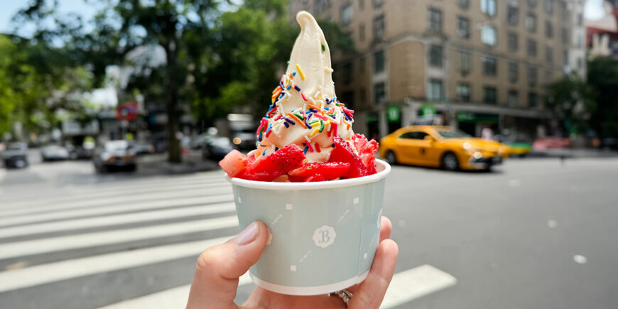 A hand holds a cup of vanilla frozen yogurt topped with fresh strawberries and rainbow sprinkles on a sunny city street, with a crosswalk and yellow taxi blurred in the background.