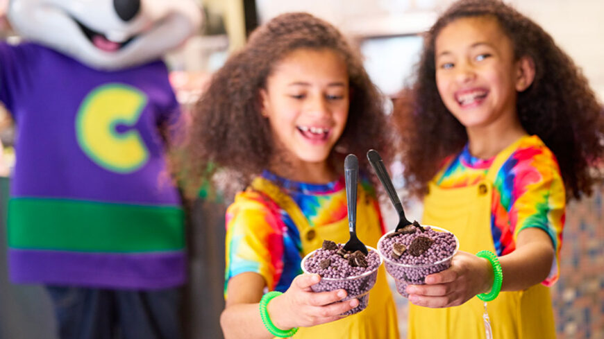 Two smiling children hold cups of purple frozen dessert topped with cookie crumbles and spoons, while a costumed mascot stands blurred in the background.