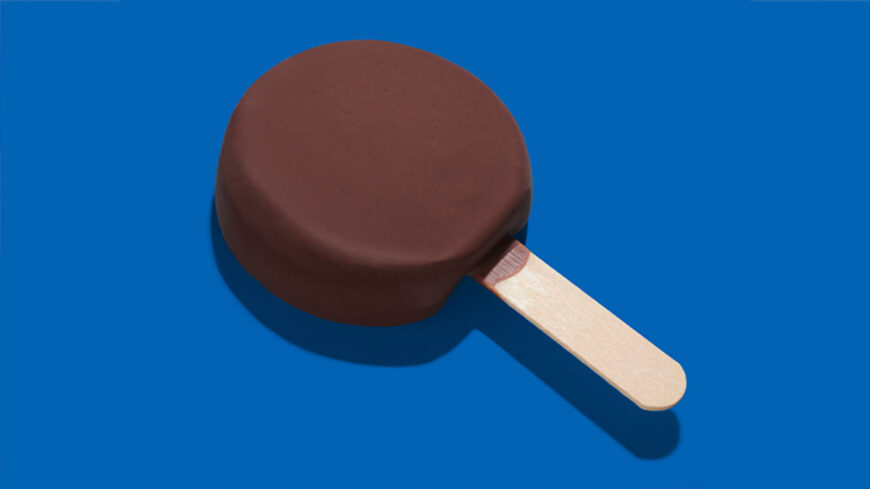 Chocolate-coated ice cream bar on a wooden stick against a solid blue background.