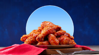 Platter piled high with glazed chicken wings on a wooden board, with a red cloth and blue background.