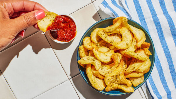 A hand dips a seasoned potato wedge into a bowl of marinara sauce next to a blue bowl filled with crispy potato wedges on a tiled countertop.