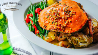 Whole roasted Dungeness crab topped with garlic herb butter, served with green beans, roasted potatoes, peppers, and lemon on a white plate.