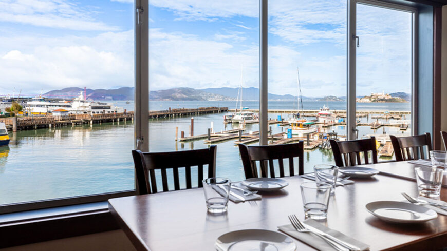 Dining room with tables set beside large windows overlooking a marina, boats on the water, and a view of San Francisco Bay under a blue sky.