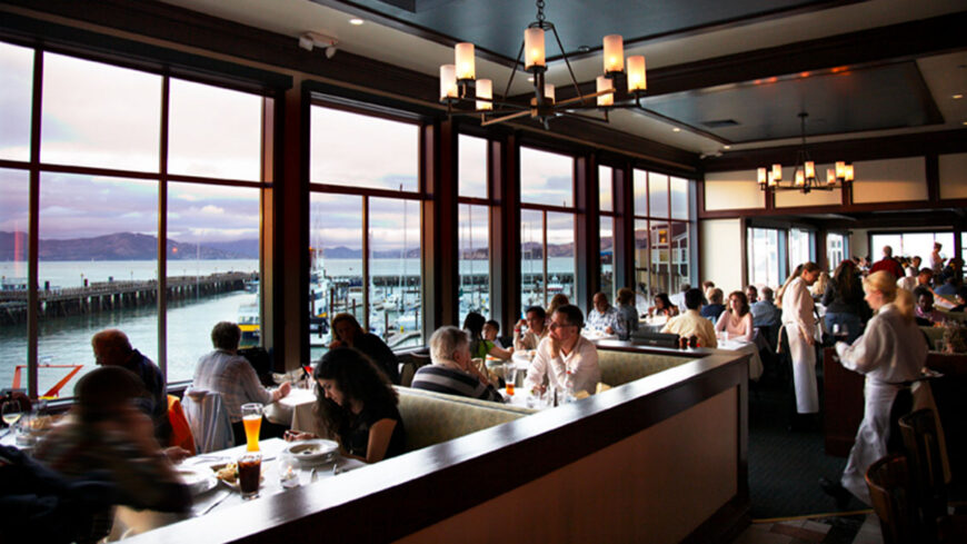 Spacious waterfront restaurant dining room with guests enjoying meals at white tablecloth tables beside large windows overlooking a marina and bay at sunset.