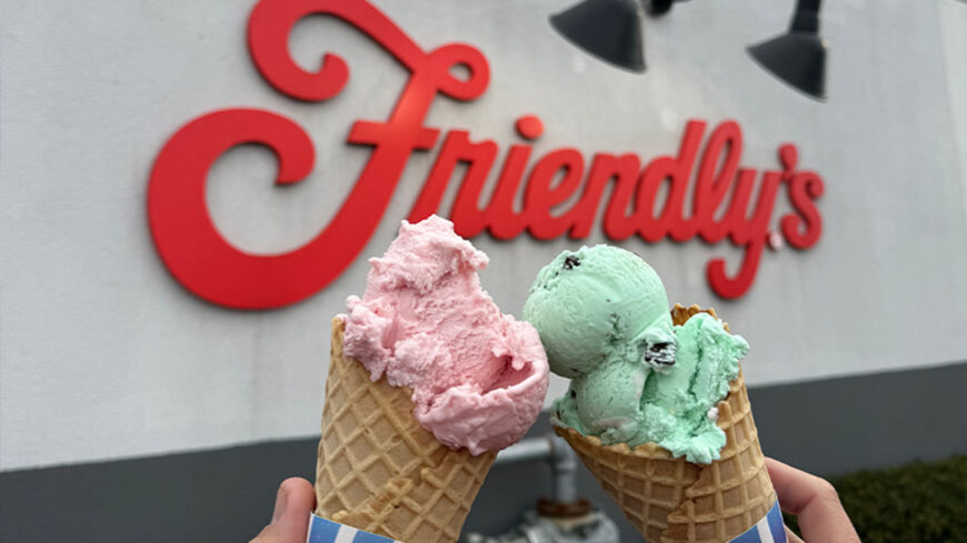 Two waffle cones filled with pink and mint green ice cream are held up in front of a large red Friendly's sign outside the shop.