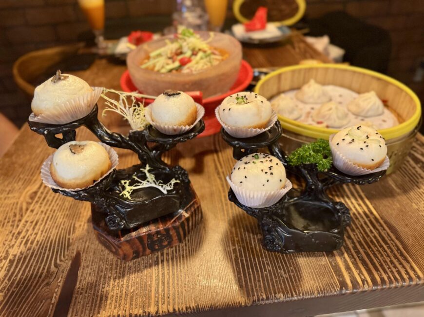 A wooden restaurant table with two decorative black tree-shaped stands displaying six steamed bao buns in white paper liners. Some buns are topped with black sesame seeds or small garnishes. In the background, slightly out of focus, are a bamboo steamer filled with dumplings, a large bowl of salad, two orange-colored drinks, and slices of red velvet cake, creating the setting of a shared dim sum meal.
