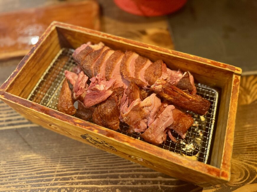 A wooden tray filled with sliced smoked duck, crispy duck skin, and tender shredded duck meat, served on a wire rack over a rustic wooden table.
