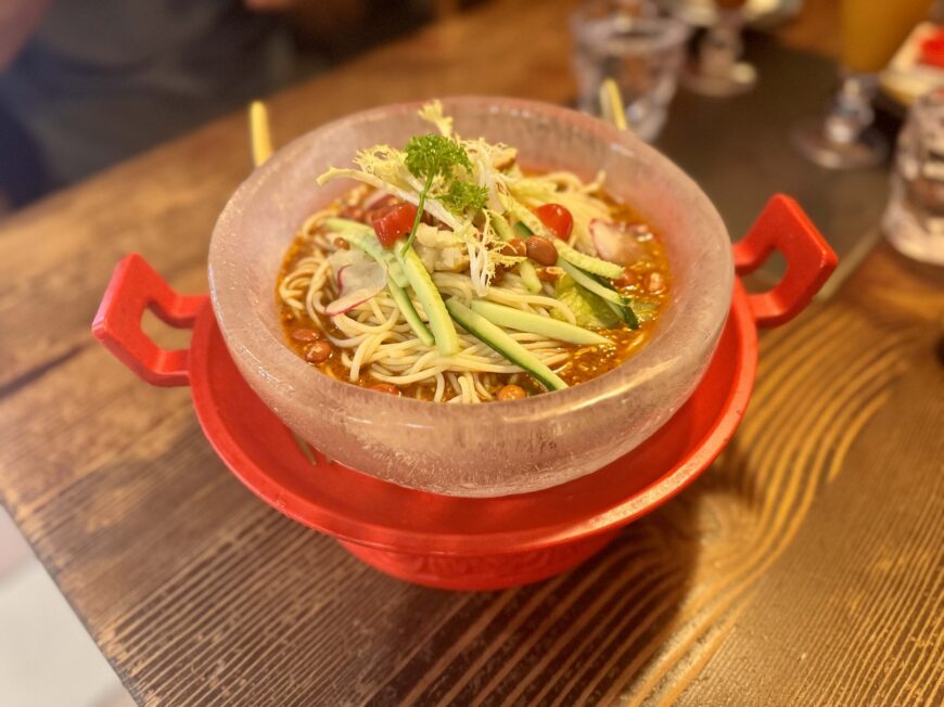 A bowl of chilled noodles in a spicy sauce, topped with cucumber, cherry tomatoes, radish slices, peanuts, and fresh parsley, served in an ice bowl on a red stand.