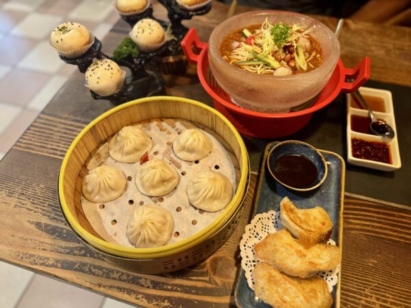 A table spread featuring steamed soup dumplings, pan-fried dumplings, chilled spicy noodles, steamed buns, dipping sauces, and a smoked duck platter, arranged on a rustic wooden table.