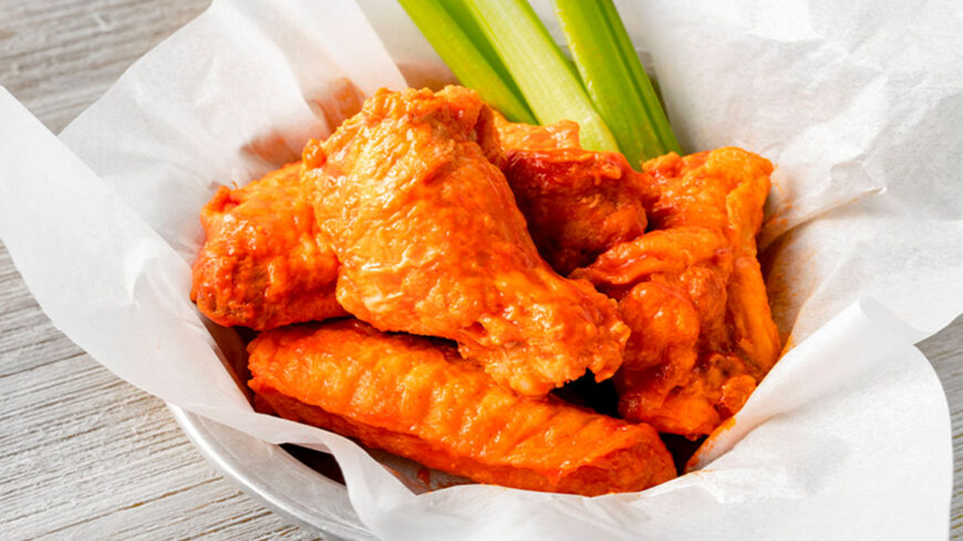 Basket of Buffalo chicken wings covered in bright orange hot sauce, served with fresh celery sticks.