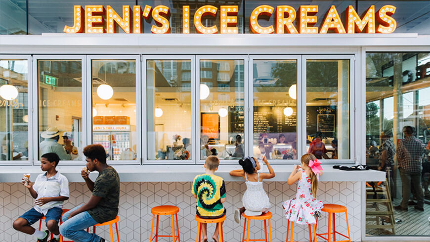 Children sit on orange stools enjoying ice cream outside a Jeni's Ice Cream shop with large illuminated signage above the windows.