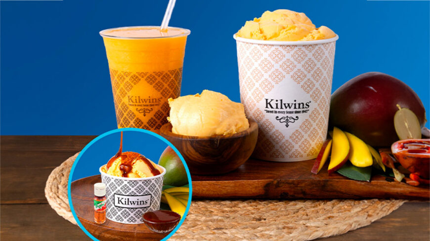 A Kilwins mango-themed display featuring a mango shake, cups of mango ice cream, fresh mangoes, and a caramel-topped sundae on a wooden table against a blue background.