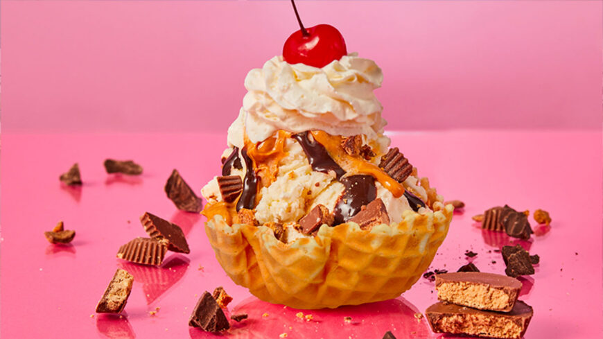 A waffle bowl filled with vanilla ice cream, topped with peanut butter and chocolate sauce, whipped cream, a cherry, and chunks of chocolate candy on a pink background.