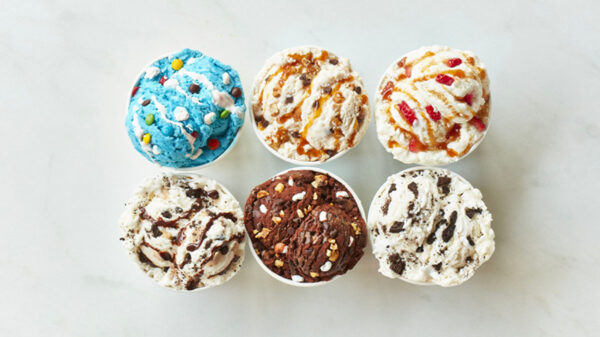 Six cups of assorted ice cream flavors viewed from above, featuring colorful mix-ins, caramel drizzle, chocolate, cookies, and candy pieces on a white marble surface.