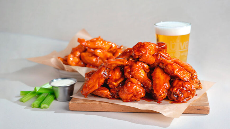 A large pile of glossy, sauce-coated chicken wings served on a wooden board with celery sticks, ranch dressing, and a glass of beer, with another basket of wings in the background.