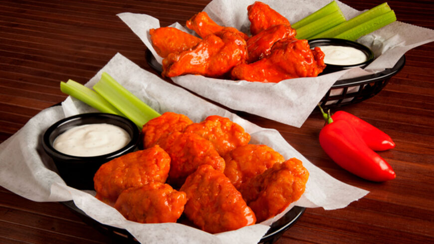 Two baskets of buffalo chicken wings served with celery sticks and ranch dipping sauce on a wooden table, with red chili peppers placed beside the baskets.