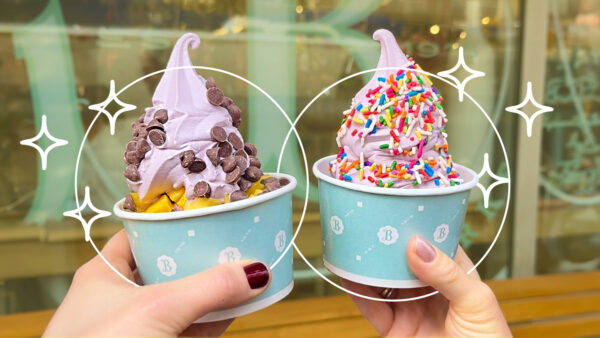 Two hands hold cups of purple frozen yogurt outside a storefront—one topped with chocolate chips and mango pieces, the other with rainbow sprinkles—with decorative sparkle graphics surrounding the desserts.